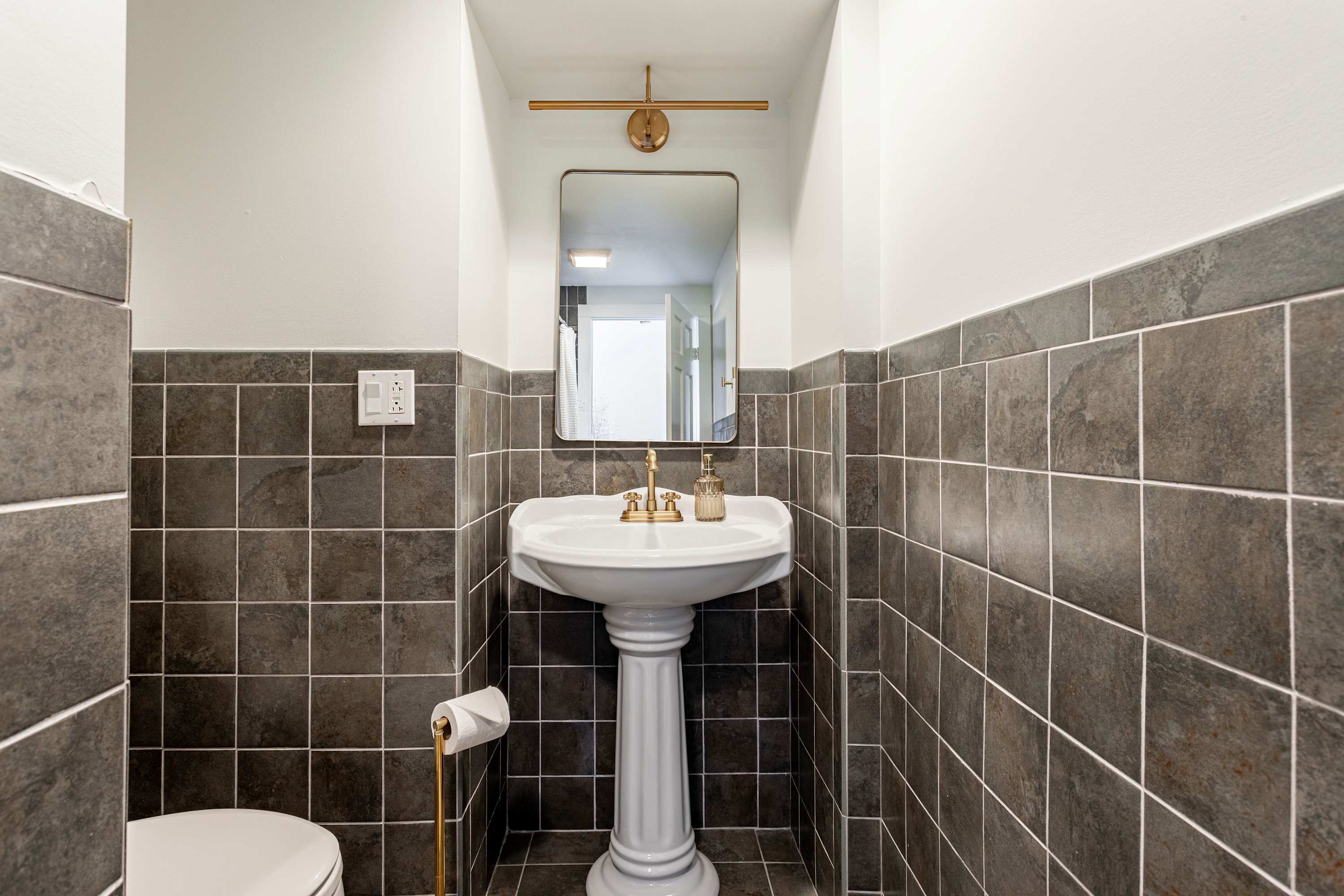 A compact bathroom featuring a white pedestal sink with a gold-toned faucet and a rectangular mirror with a gold light fixture above. Dark gray square tiles cover the lower half of the walls and the floor.