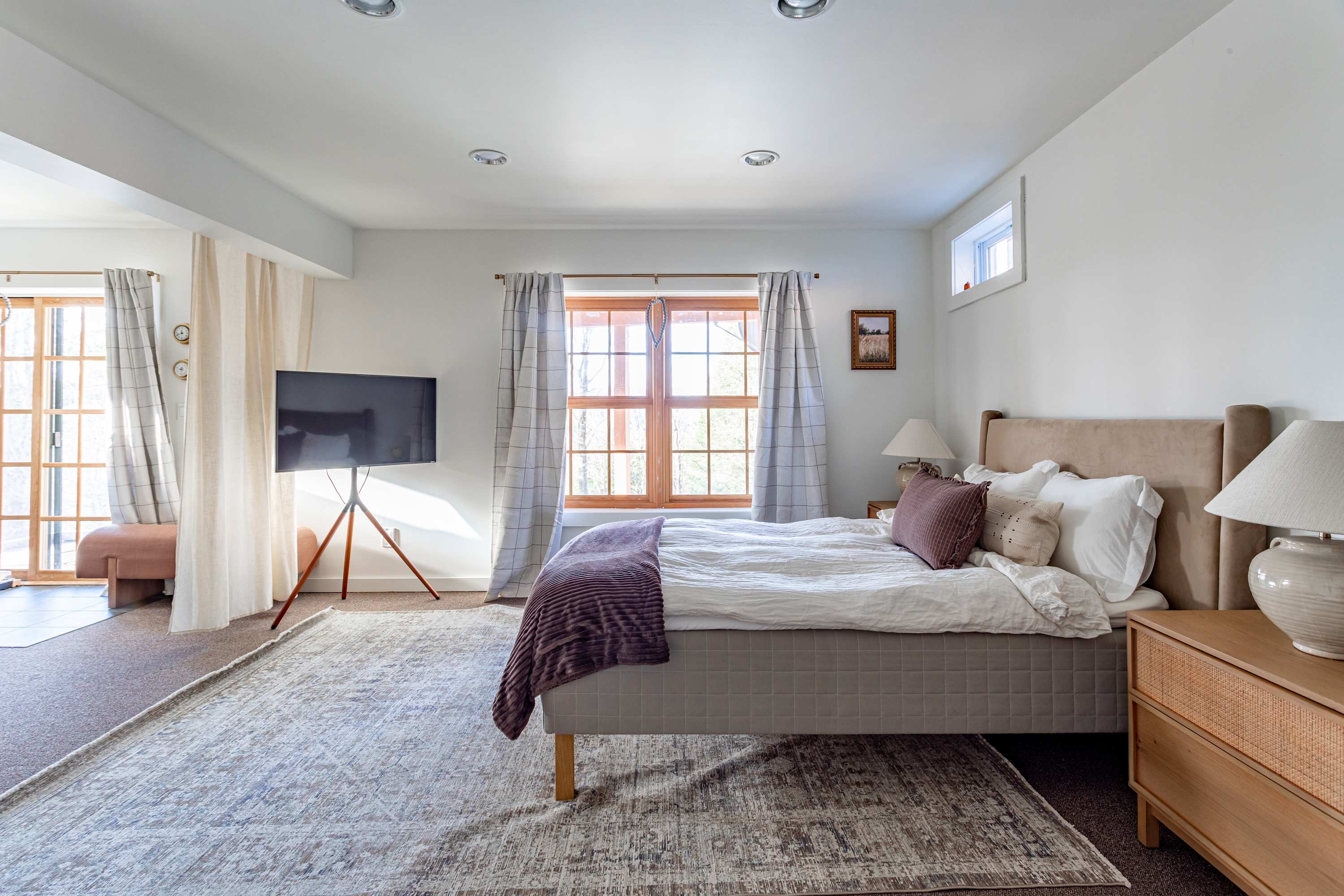 A modern bedroom with white walls and dark carpet, featuring a queen-sized bed with a beige upholstered headboard, two wooden nightstands with lamps, and a large window with plaid curtains. A TV on a stand is visible to the left.