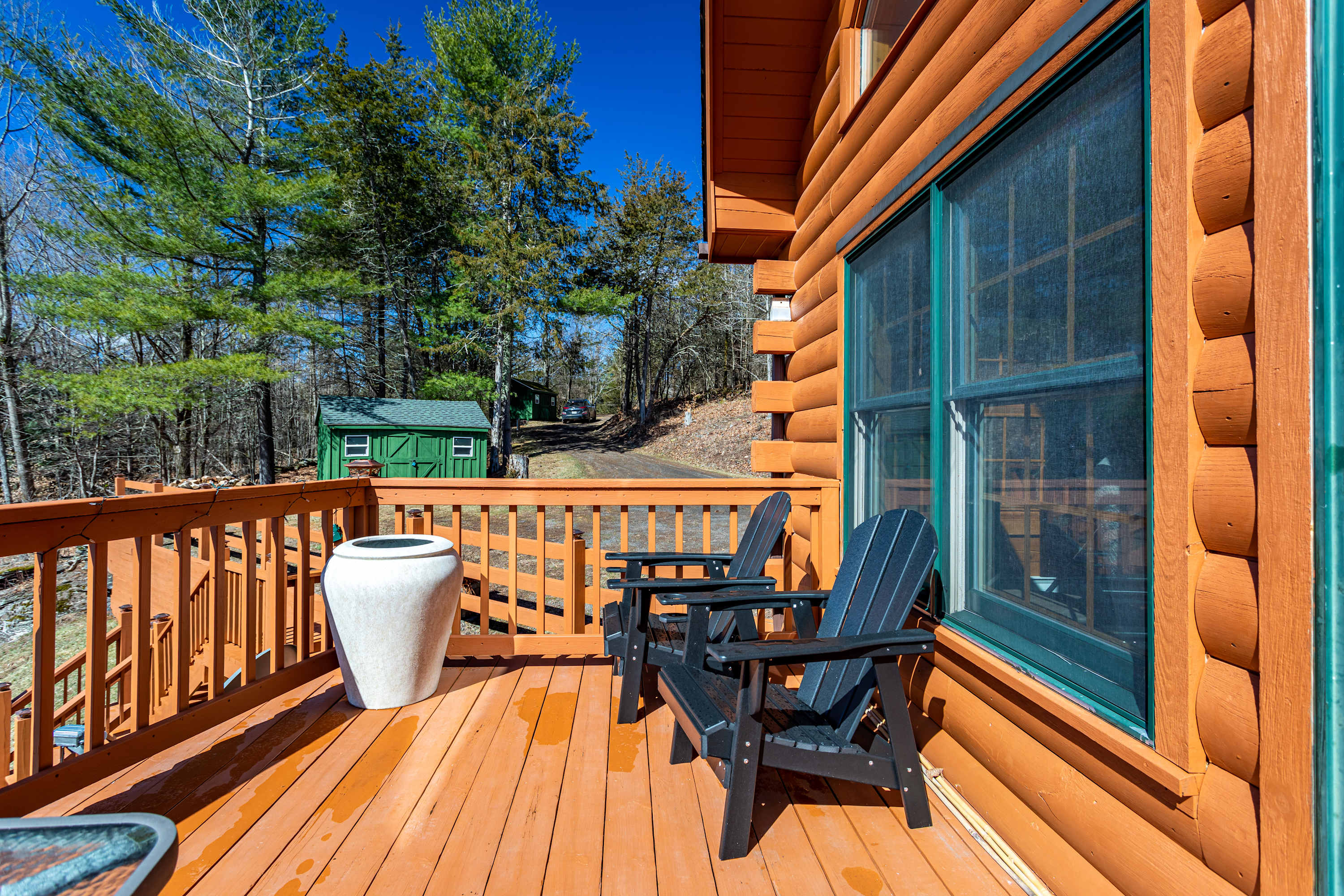 A section of a wooden deck with two black Adirondack chairs and a large white planter, offering a view of a gravel driveway and a green shed in the distance, surrounded by trees.