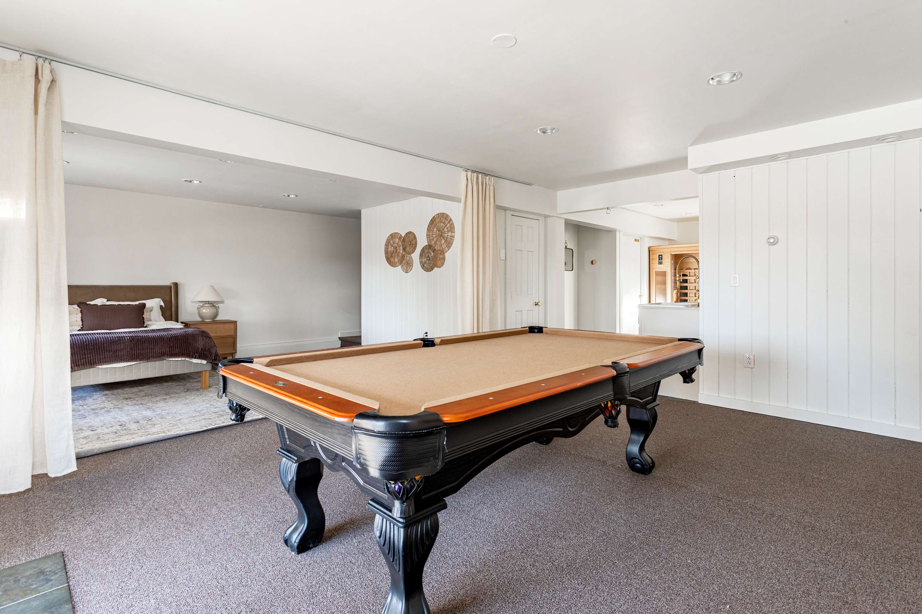 A spacious recreation area featuring a pool table with a tan felt surface, dark carpet, and white walls. Part of a bedroom with a queen bed and a window is visible in the background.