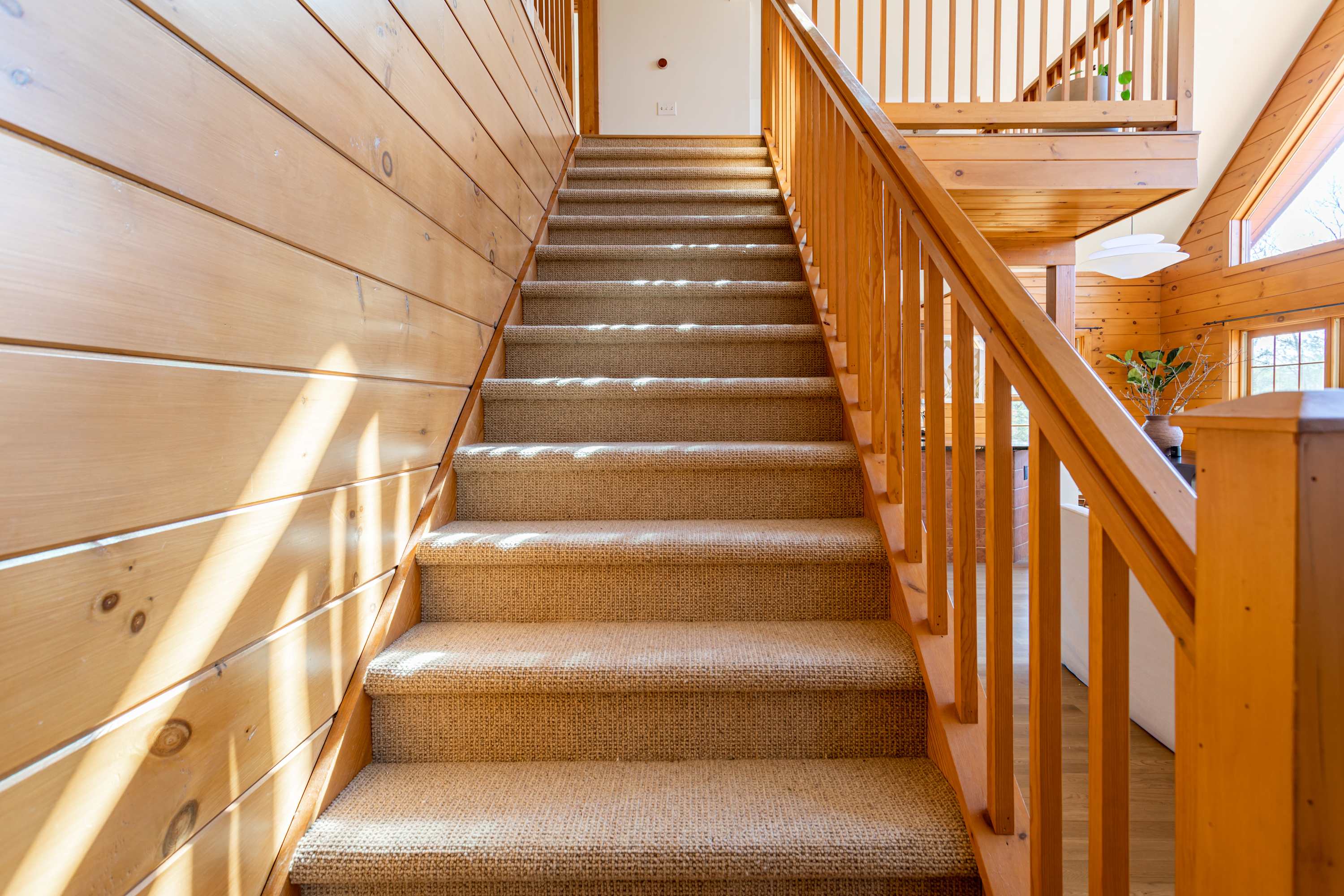 A carpeted wooden staircase with a natural wood railing, leading to the upper floor. Sunlight streams through a nearby window, highlighting the wood paneling.