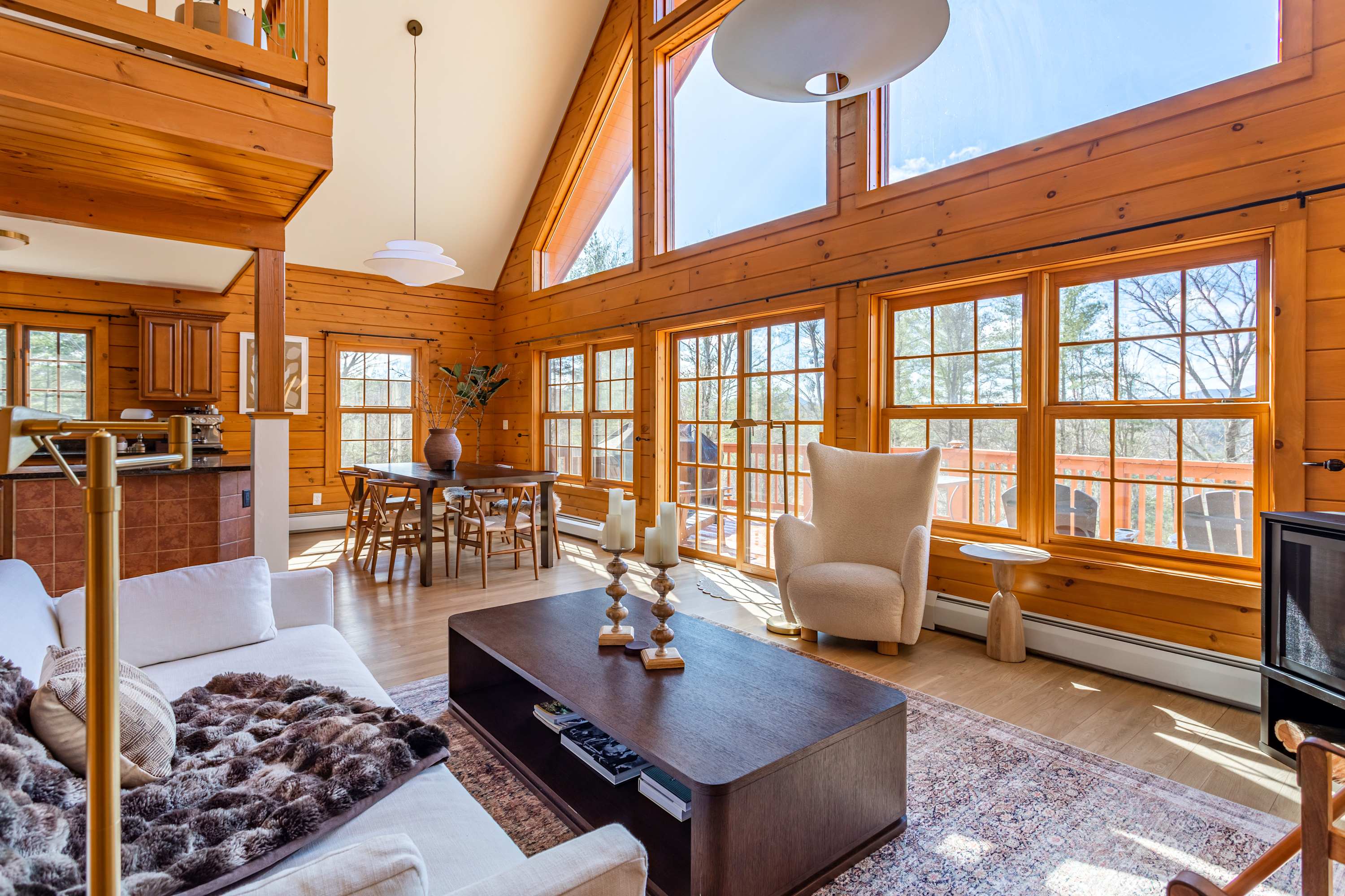 A bright, open-concept living space with light wood paneling, a white sofa, dark wood coffee table, and large windows offering natural light and views. A dining area is visible in the background.