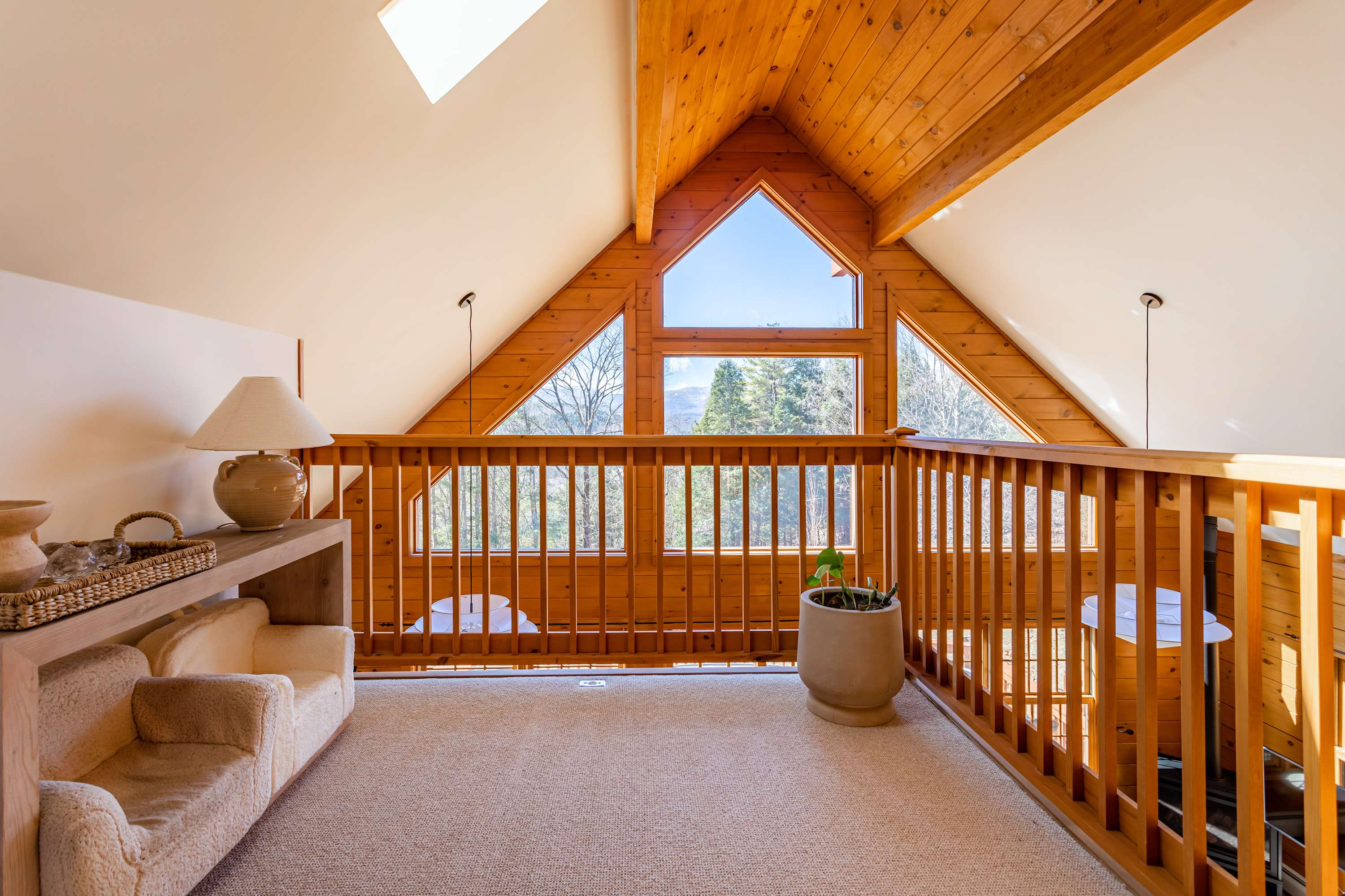 Cozy carpeted loft area overlooking the main living space, featuring a small armchair, a lamp, and a wooden railing, with a triangular window offering views of the surrounding trees.