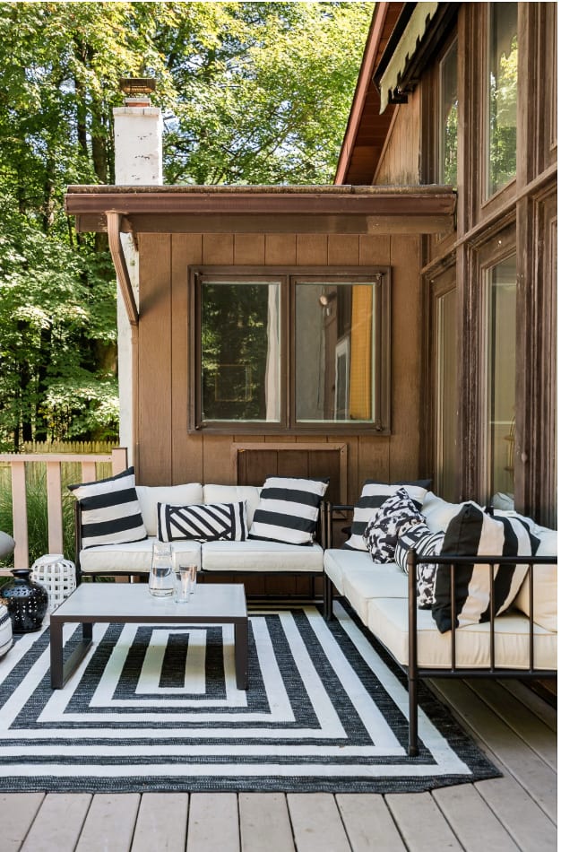 A cozy outdoor seating area on a wooden deck features a black and white striped rug, a dark metal coffee table, and two white sofas with black and white striped cushions. The house exterior with brown siding and large windows is visible behind the furniture.