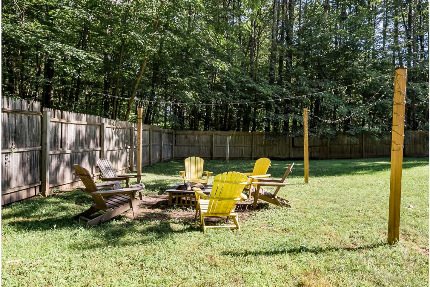 A fire pit area in a grassy backyard is encircled by several Adirondack chairs, some wooden and some bright yellow, with string lights strung between wooden posts overhead. A wooden fence and dense trees create a private, natural setting.