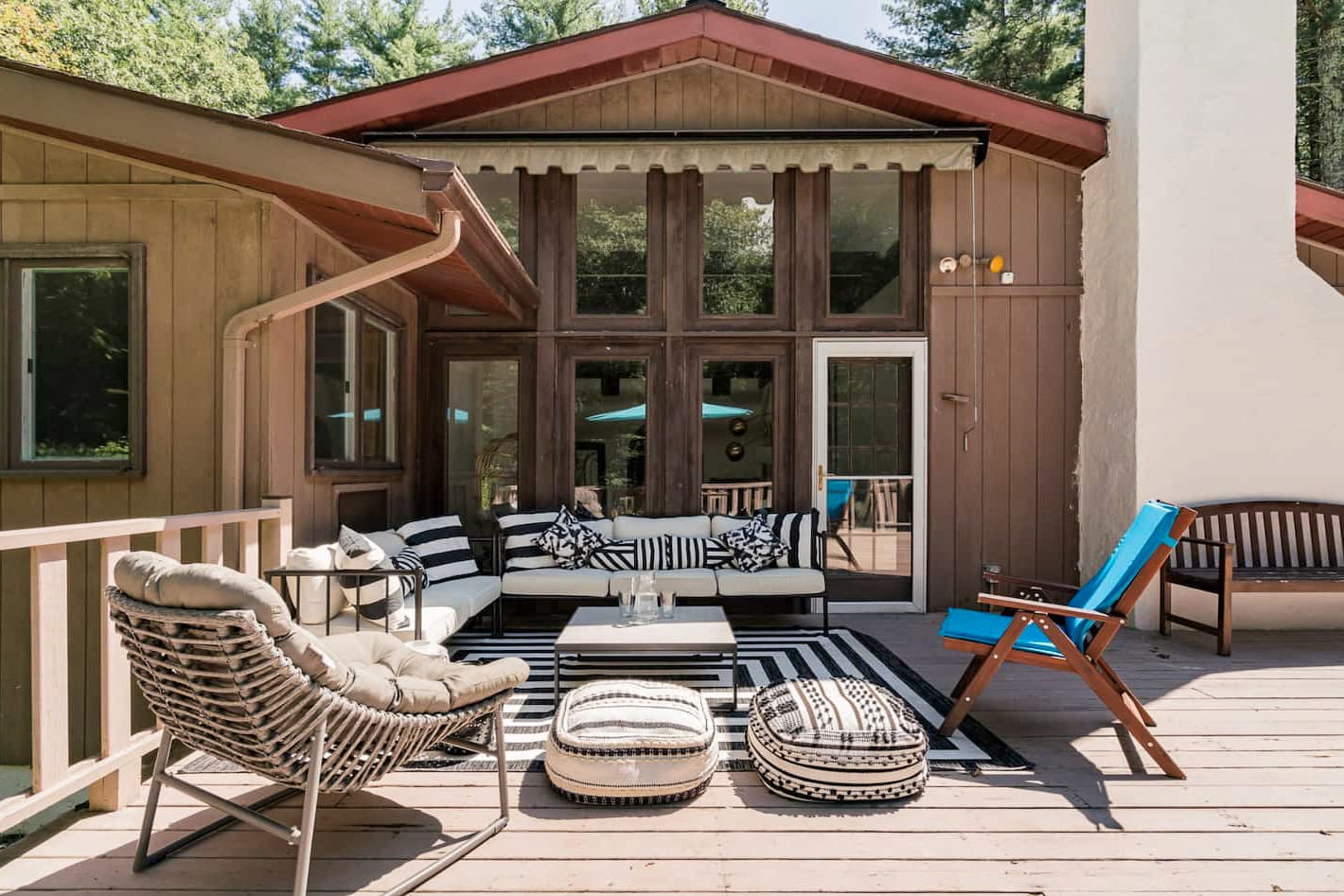 A spacious wooden deck features a modern outdoor sectional with black and white striped pillows, a coffee table, and a wicker chair, set against a brown-sided house with large windows.