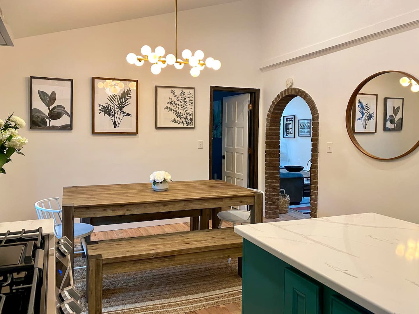 A modern dining area features a large wooden dining table with a bench and white chairs, illuminated by a contemporary gold chandelier. The space connects to a kitchen island with a white countertop and an arched doorway leading to another room.