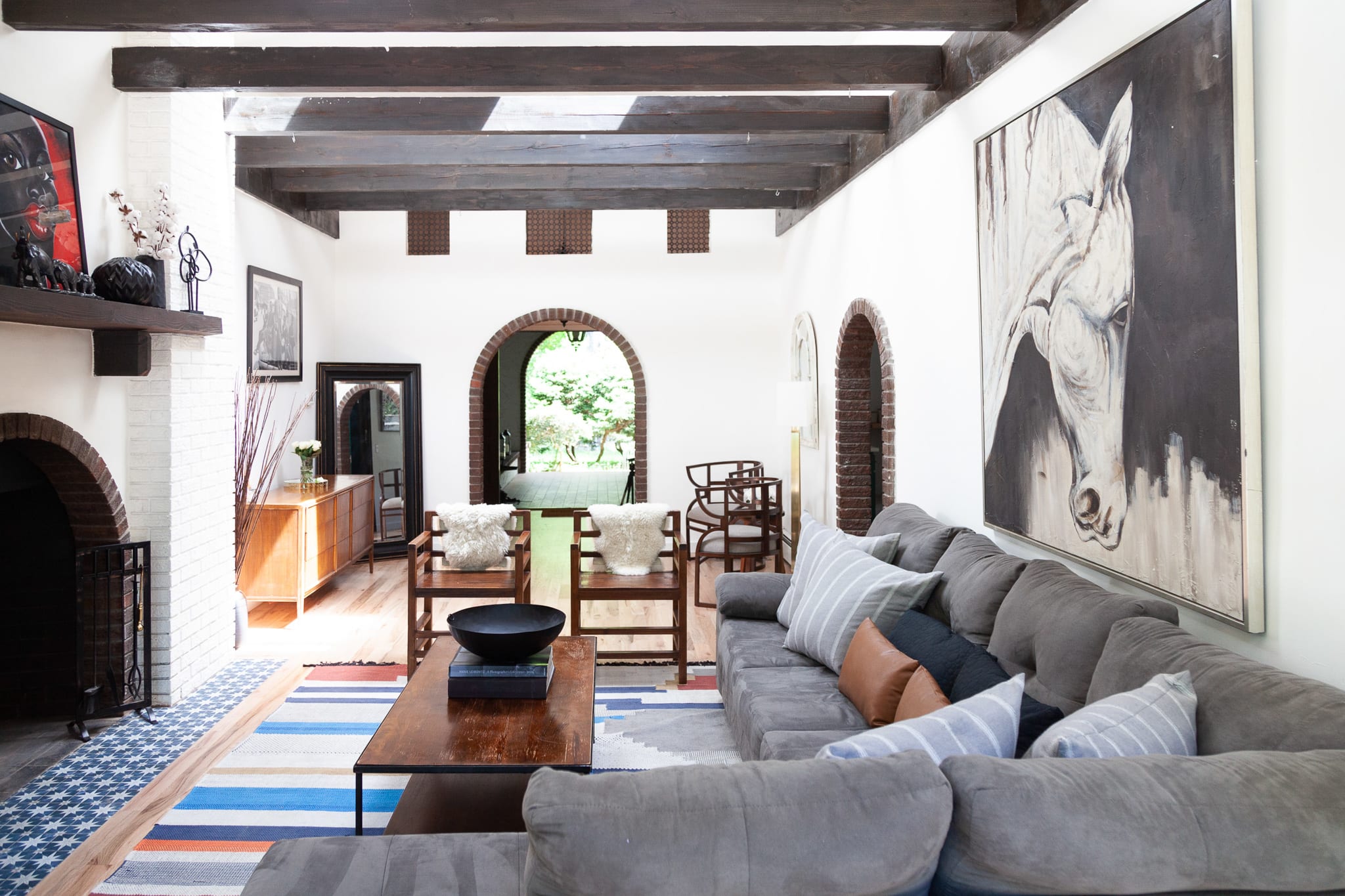 A spacious living room with exposed dark wood ceiling beams features a large grey sectional sofa, a wooden coffee table, and a striking horse painting on the white wall, with brick archways leading to other rooms.