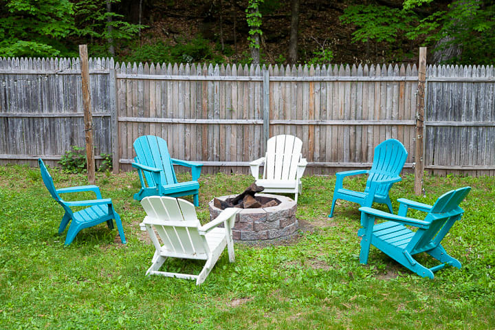 A circular stone fire pit is centered in a grassy area, surrounded by six Adirondack chairs in varying shades of blue and white. A rustic wooden fence forms the backdrop, with dense green trees beyond.