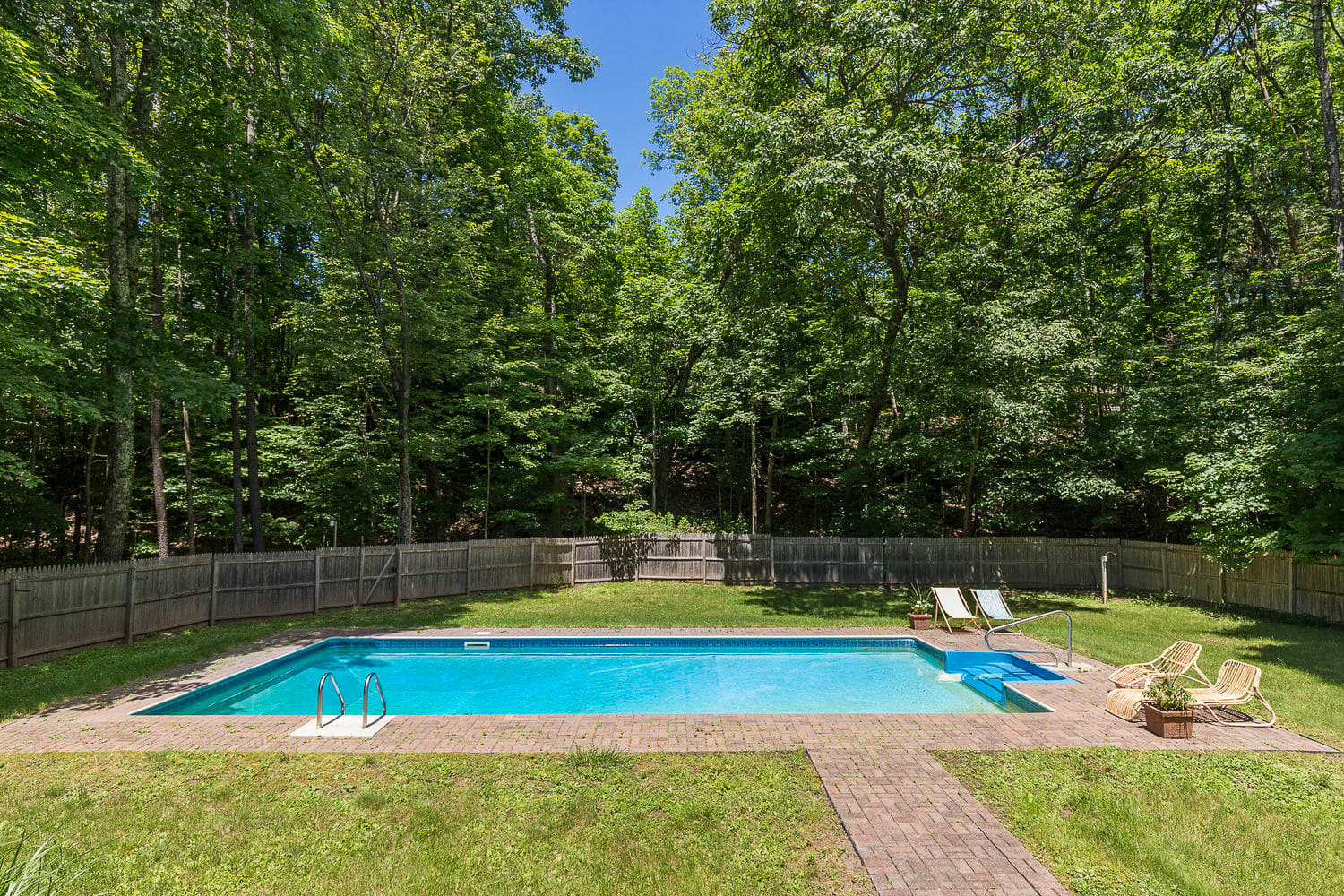 An inviting rectangular swimming pool with blue tiling is set in a grassy backyard, surrounded by a brick patio and a wooden fence, with lush green trees providing a natural backdrop. Two white lounge chairs sit on the far side of the pool.