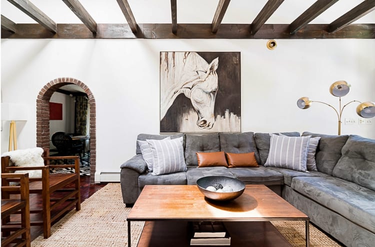 A comfortable living room features a large grey sectional sofa, a wooden coffee table, and a prominent horse painting on the wall, with dark exposed ceiling beams and a brick archway leading to another room.