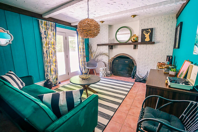 A vibrant living room with teal paneled walls and a white brick fireplace, featuring a black and white striped rug and a green velvet sofa. A round coffee table and a woven pendant light complete the eclectic decor.