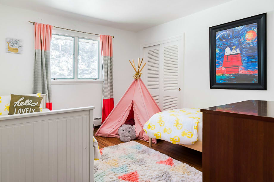 A playful children's bedroom with white walls and dark wood floors, featuring two twin beds with yellow monkey-patterned bedding and a pink teepee. A colorful rug and a large framed artwork add to the vibrant decor.