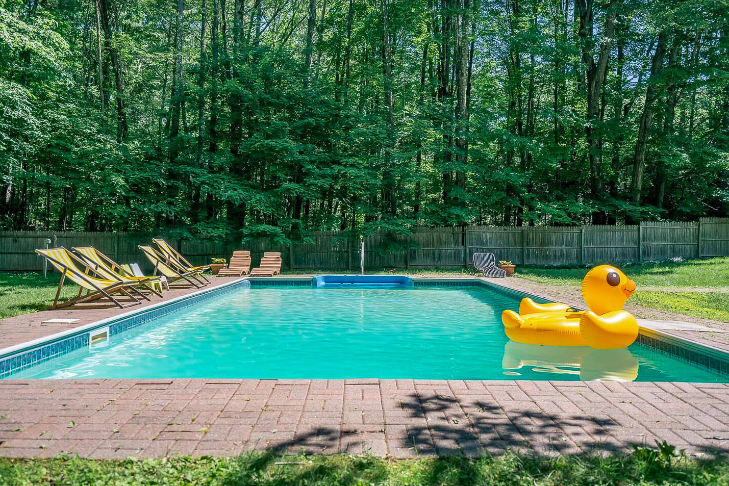 A large rectangular outdoor swimming pool surrounded by a brick patio and several yellow and white striped lounge chairs. The pool is set against a lush green backdrop of dense trees, with a large yellow duck float in the water.
