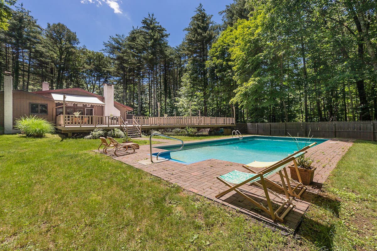 A rectangular swimming pool with blue water is surrounded by a brick patio and a green lawn, with a large wooden deck and a house visible in the background. Tall pine trees and other lush foliage frame the scene under a bright sky.
