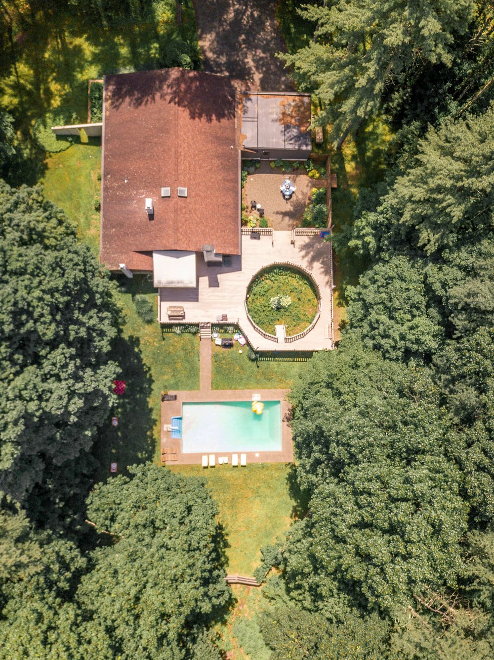 An aerial view shows a large house with a brown roof surrounded by dense green trees, featuring a rectangular swimming pool in the foreground and a circular garden area on a multi-level deck. The property is nestled within a lush, wooded landscape.