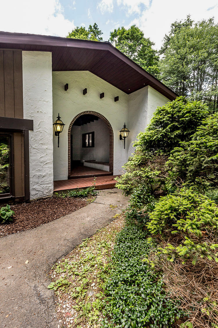 The front entrance of the house features a white stucco facade with a dark brown arched entryway, flanked by two traditional black lanterns. A paved path leads to the entrance, surrounded by lush green shrubs and ground cover.