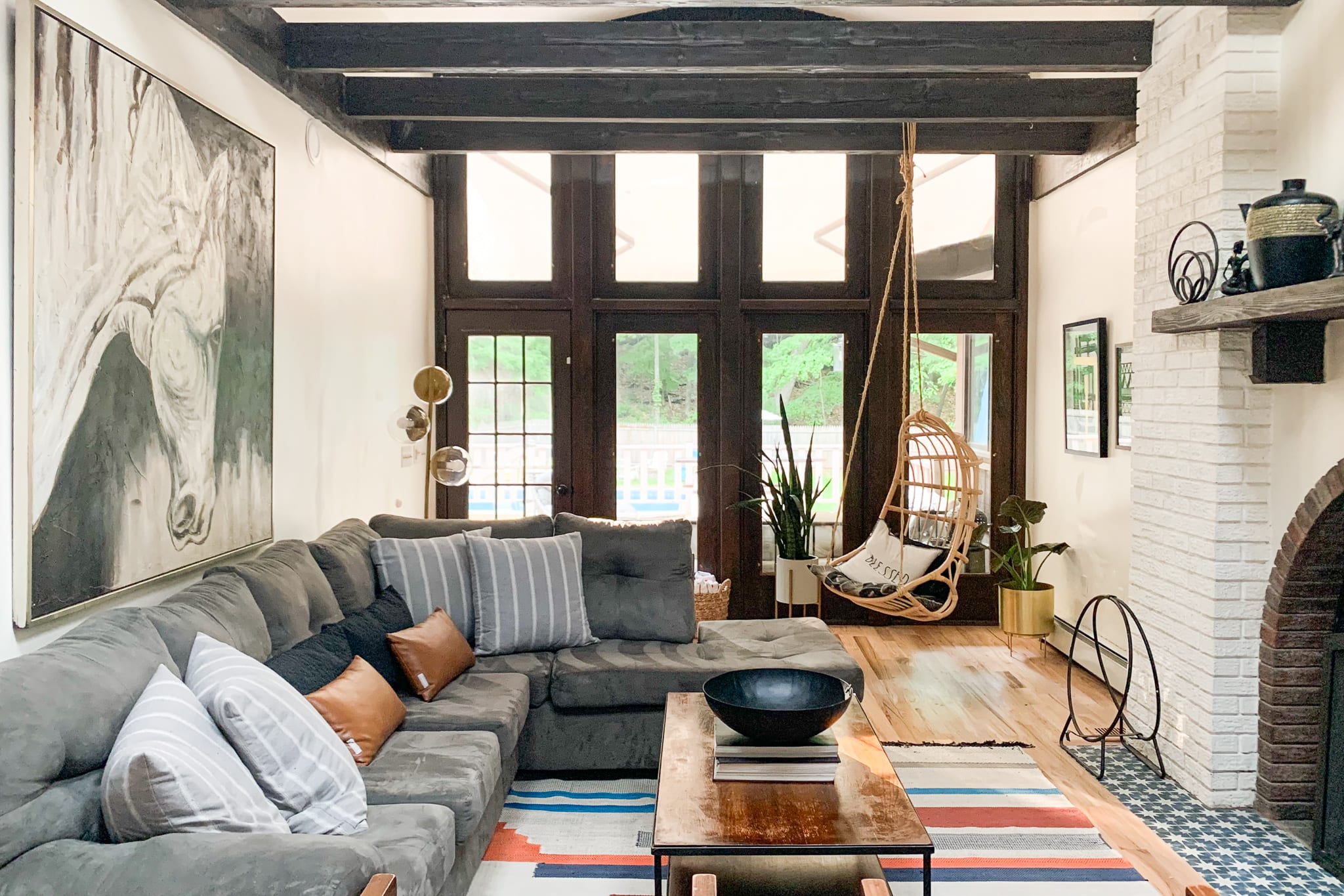 This living room features a large grey sectional sofa, a wooden coffee table, and a white brick fireplace, with dark exposed ceiling beams and large glass doors leading to the outside.