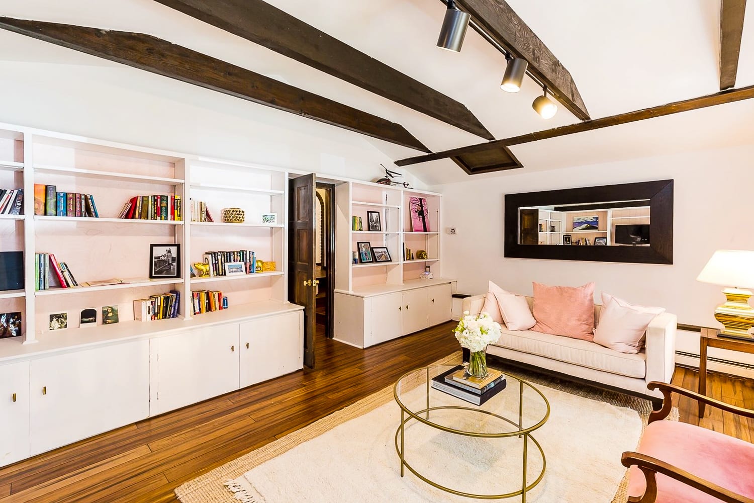 A cozy living space features extensive white built-in bookshelves filled with books and decor, a light-colored sofa with pink pillows, and a glass coffee table, all under a vaulted ceiling with dark exposed beams.