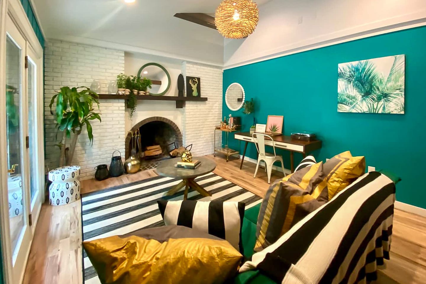 A bright and eclectic living room with a white brick fireplace and vibrant teal accent wall, furnished with a green sectional sofa and a black and white striped rug. A desk area with a white chair and various plants adds functionality and greenery.