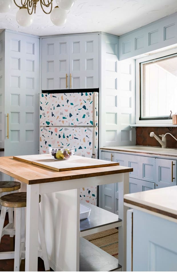 A unique kitchen features light blue paneled cabinetry and a white refrigerator with a colorful terrazzo-like pattern. A wooden island with stools provides additional workspace, and a window offers natural light.