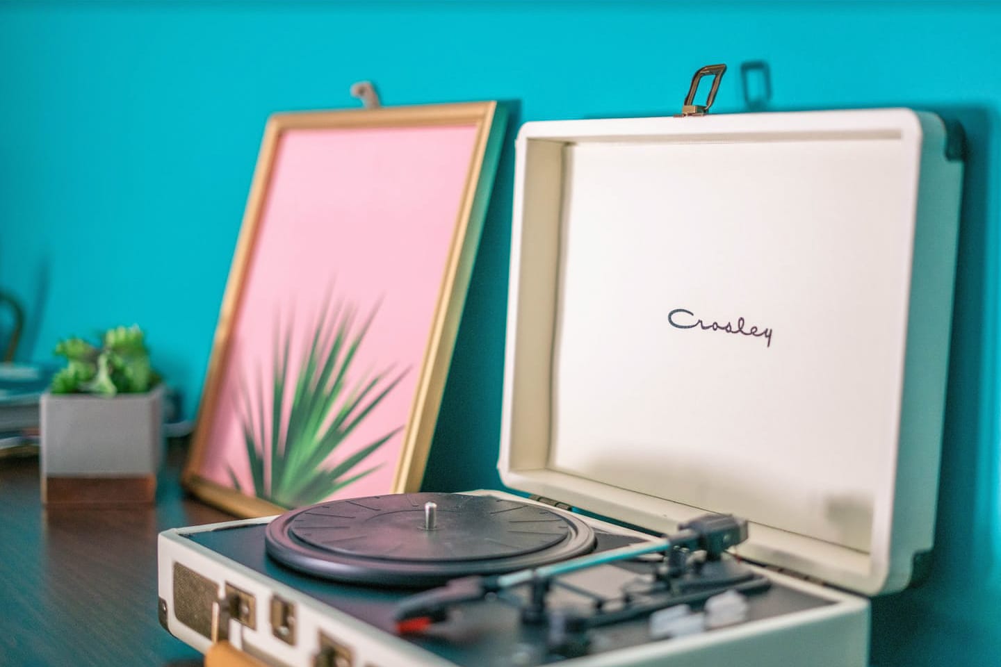 A close-up shot of a vintage-style white record player on a wooden surface, next to a framed pink and green botanical print. The vibrant teal wall serves as a colorful backdrop, adding to the room's eclectic charm.