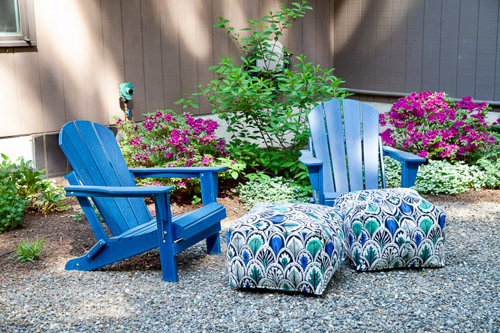Two bright blue Adirondack chairs are placed on a gravel path next to a garden bed with vibrant pink and green foliage, against the backdrop of a brown house exterior. Two patterned ottomans sit between the chairs, adding a pop of color.