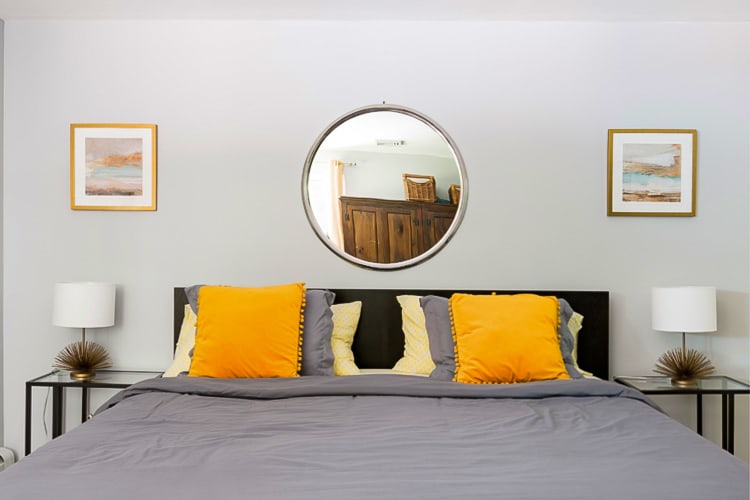 A neatly arranged bedroom with a bed featuring gray bedding and bright yellow accent pillows, flanked by two modern nightstands with lamps. A large round mirror hangs above the headboard, centered between two framed artworks.