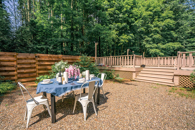 An outdoor dining area on a gravel patio features a table draped with a blue and white tablecloth, set with plates and wine glasses, surrounded by four white metal chairs. A multi-level wooden deck is visible in the background, with lush green trees beyond.