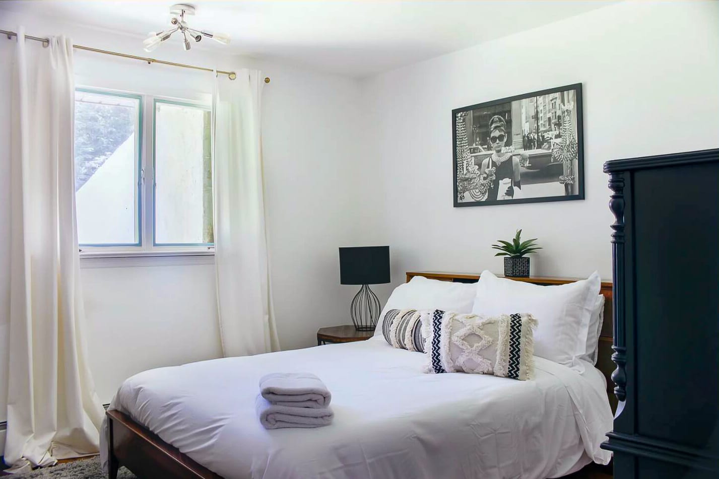 A cozy bedroom with white walls and a queen-sized bed dressed in white linens and decorative pillows. A black-framed artwork hangs above the bed, and a dark nightstand with a lamp is beside it.