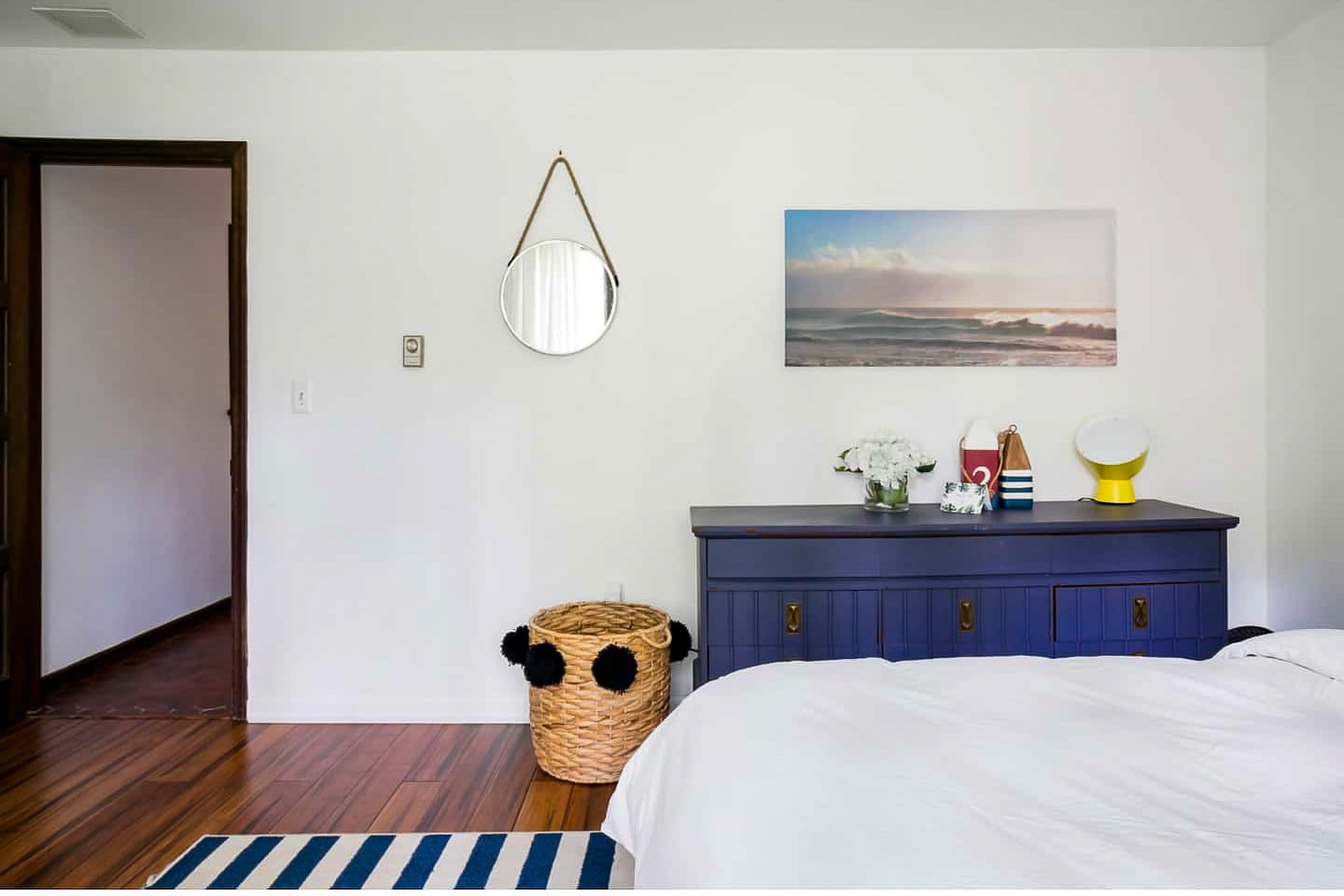 A view of a bright bedroom with white walls and dark wood floors, featuring a blue dresser with a framed ocean print above it. A round mirror hangs on the wall, and a woven basket sits beside the dresser.