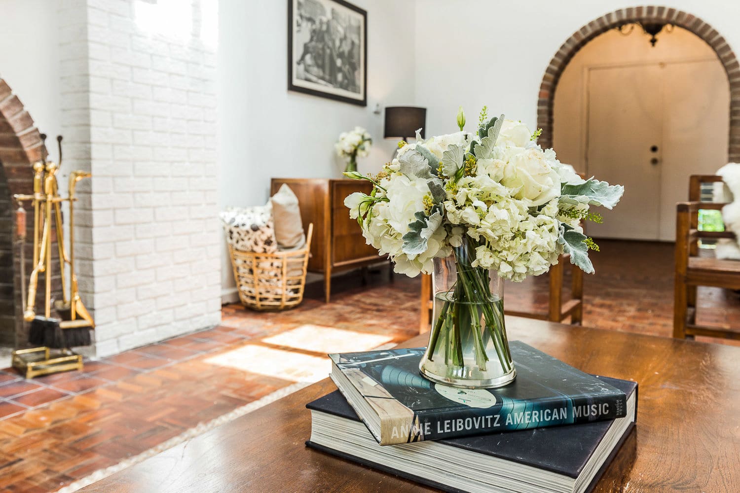 A bright living space with dark wood parquet flooring and white walls, featuring a white brick fireplace and a brick archway in the background. A wooden coffee table holds a vase of white flowers and stacked books, creating a welcoming atmosphere.