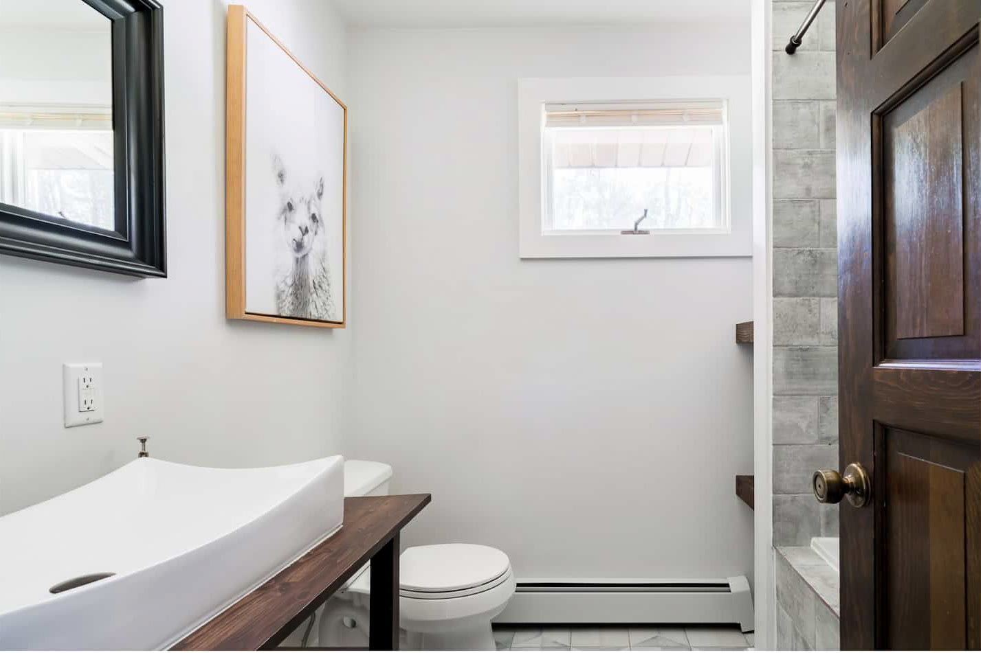 A bright bathroom with white walls, a modern vessel sink on a dark wood vanity, and a toilet. A framed llama print hangs above the toilet, and a black-framed mirror is above the sink.