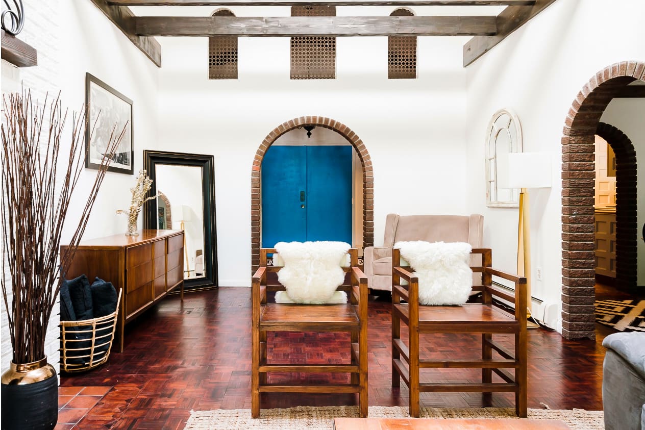 A spacious living area with dark wood parquet flooring and white walls features a prominent brick archway leading to a vibrant blue door. Two wooden chairs with white fluffy cushions and a mid-century modern credenza furnish the space.