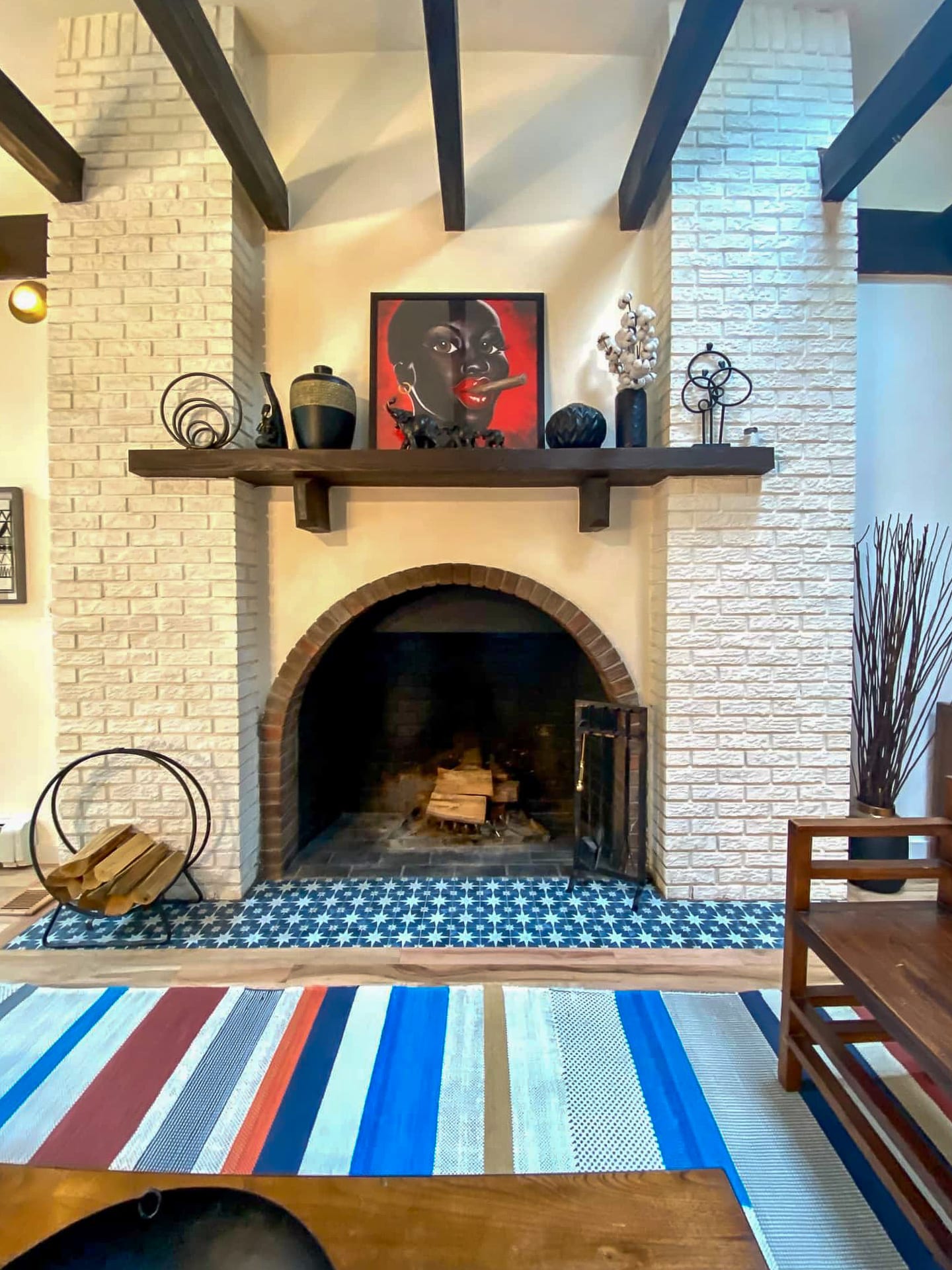 A cozy living room features a white brick fireplace with a dark wood mantel, an arched firebox, and a blue and white patterned tile hearth. A colorful striped rug adds a vibrant touch to the space, complemented by dark wood ceiling beams.