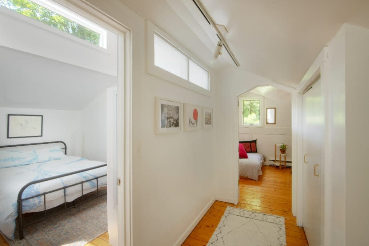 A bright hallway with light wood floors and white walls leads to a bedroom on the left, featuring a black metal bed frame with blue and white bedding and a patterned rug. Another room with a window is visible at the end of the hall.