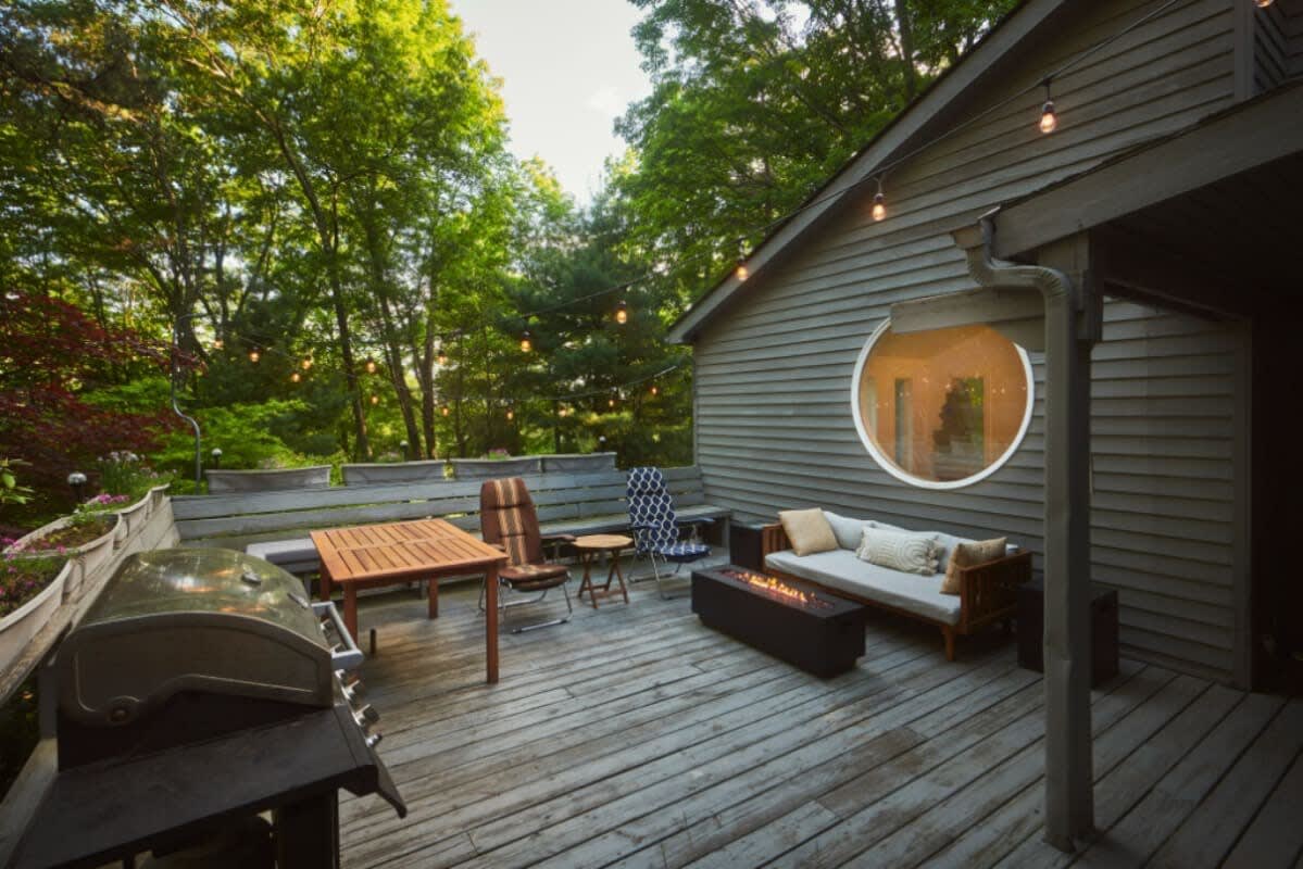 A wide wooden deck with string lights, featuring an outdoor sofa, a fire pit, a dining table, and a BBQ grill, all surrounded by lush green trees. A circular window on the dark gray house reflects the outdoor scene.