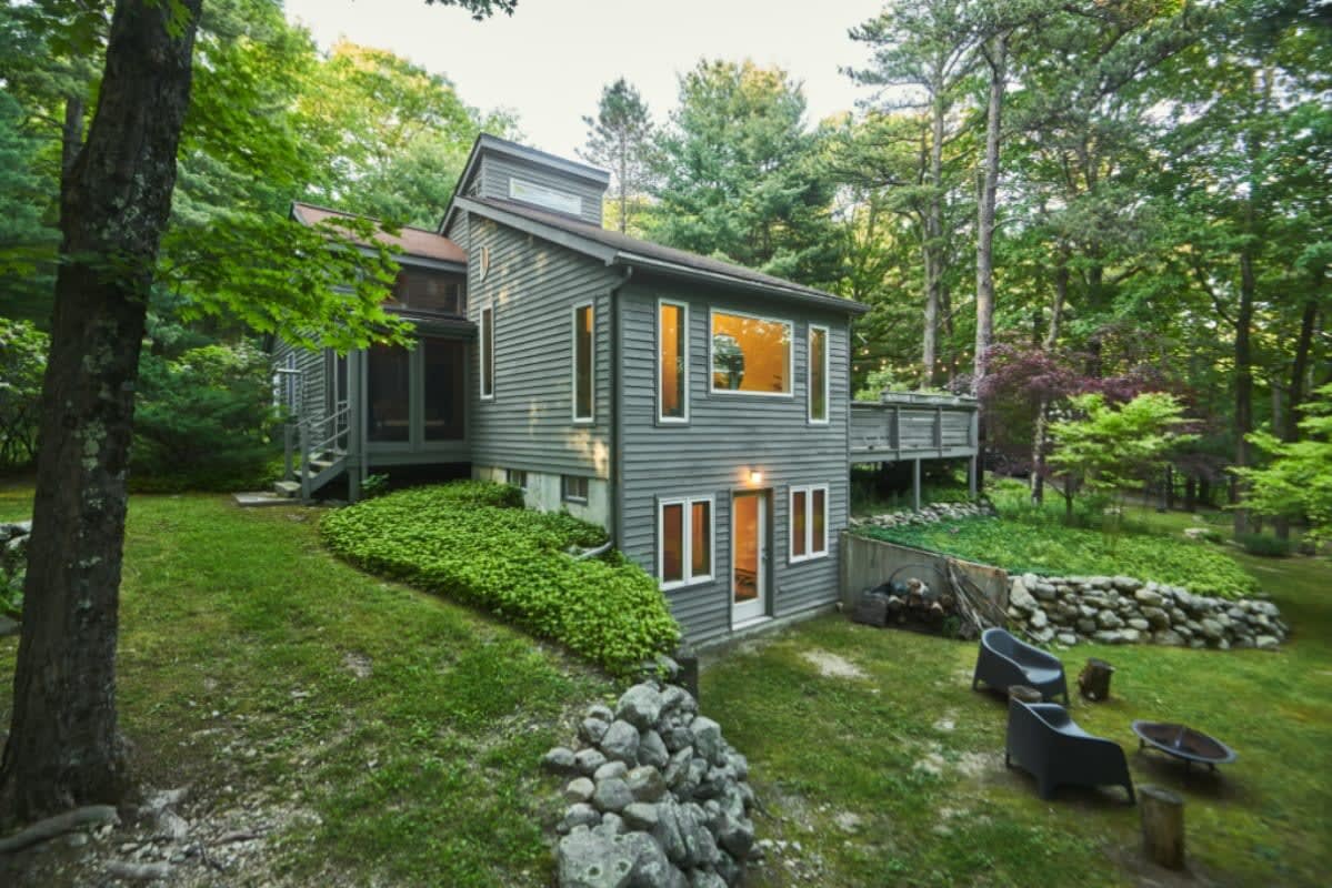 A gray-sided cabin is nestled in a lush green woodland setting, featuring multiple windows and a lower-level entrance. A small outdoor seating area with two chairs and a fire pit is visible on the grassy slope, surrounded by rock landscaping.