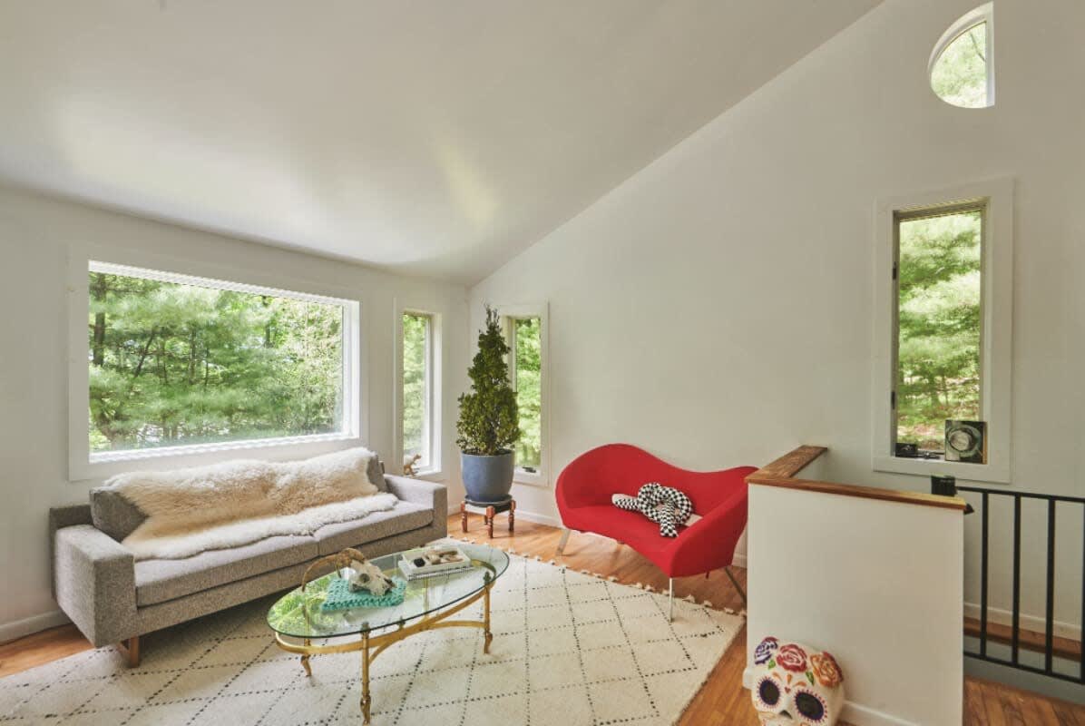 Spacious living room featuring a grey sofa with a fluffy throw, a red chaise lounge, and large windows offering expansive views of the surrounding forest. A glass coffee table sits on a patterned rug.