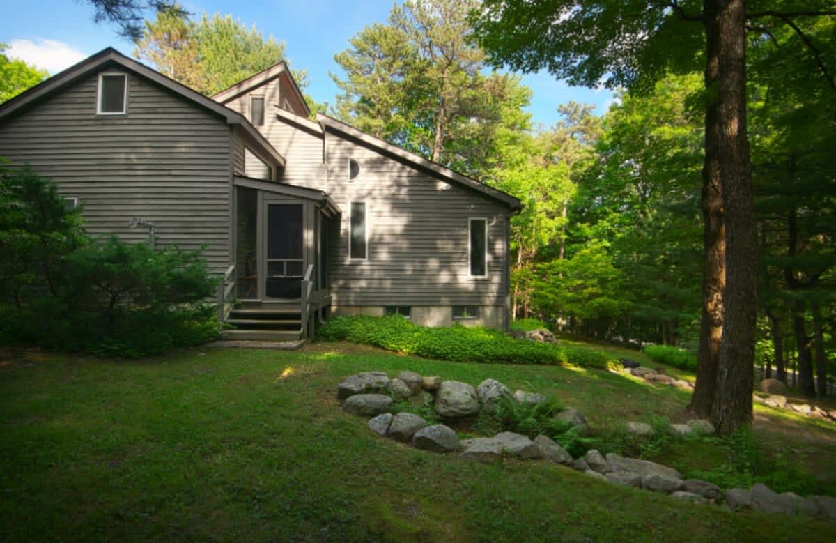 A gray-sided cabin features a screened porch and multiple windows, set on a gentle grassy slope surrounded by mature trees. Rock landscaping borders the lawn, adding to the natural aesthetic of the woodland property.