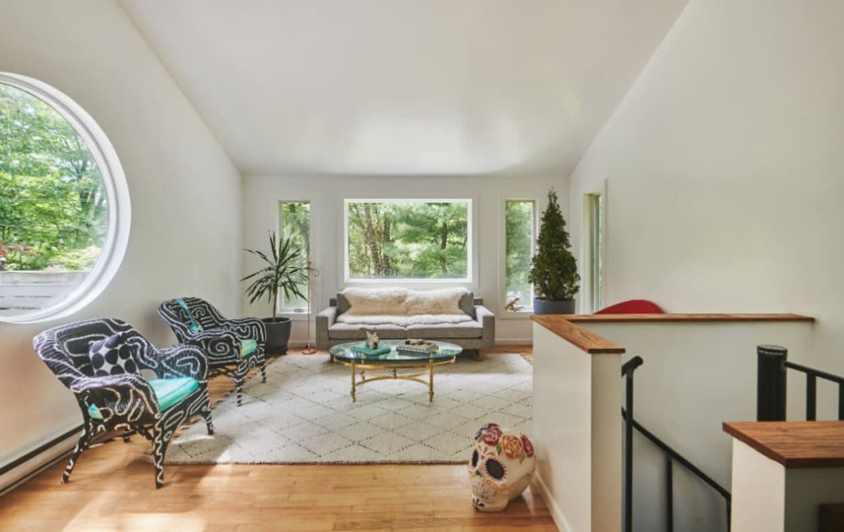 A bright living room with white walls and hardwood floors, featuring a large circular window and multiple rectangular windows offering views of the surrounding forest. A gray sofa and two black and white wicker chairs are arranged on a patterned rug.