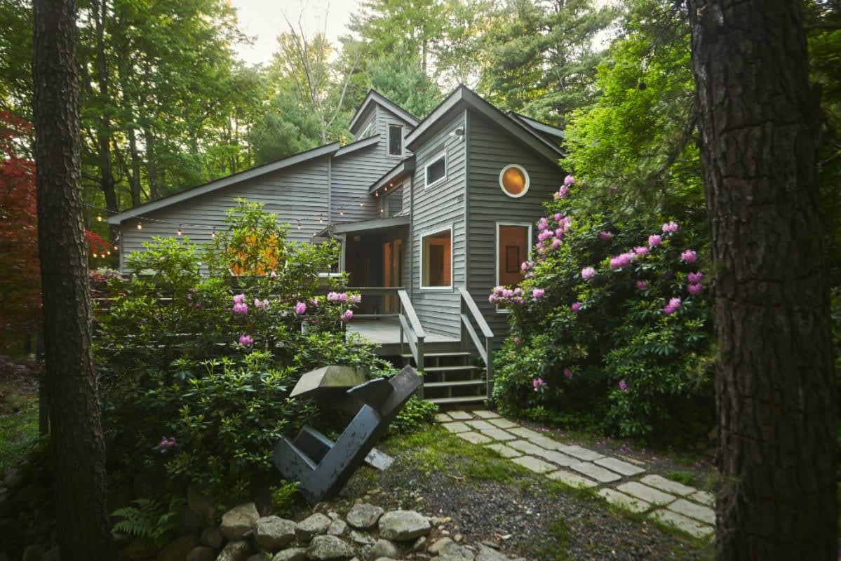 A charming gray-sided cabin is surrounded by dense green foliage and vibrant flowering bushes, with a stone path leading to a wooden staircase entrance. The house features various window shapes and is set within a natural woodland environment.