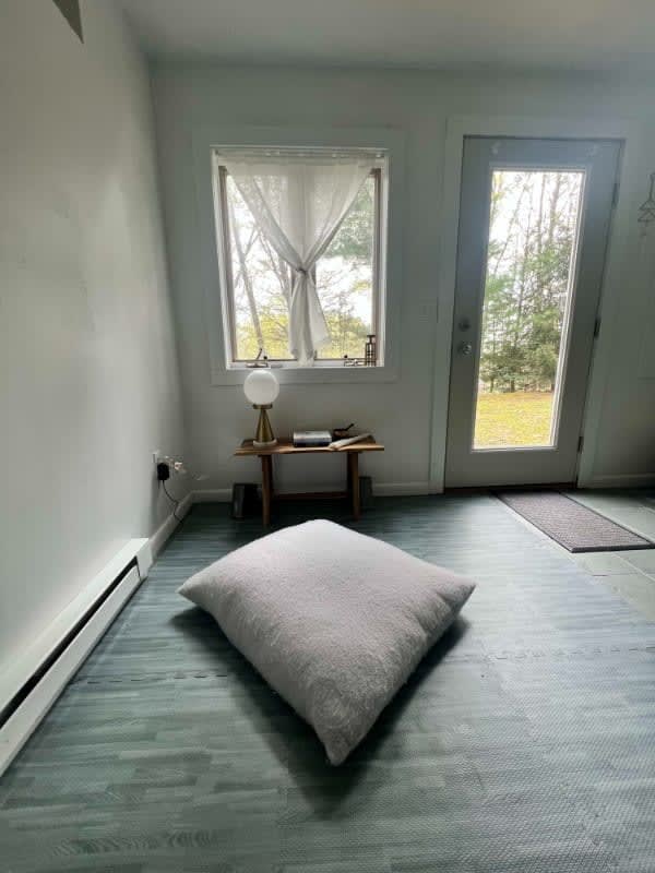 A corner of a versatile room features light gray walls, blue-toned foam floor tiles, and a large white floor cushion. A small wooden table with a lamp sits by a window with sheer curtains, next to a glass door leading outside.