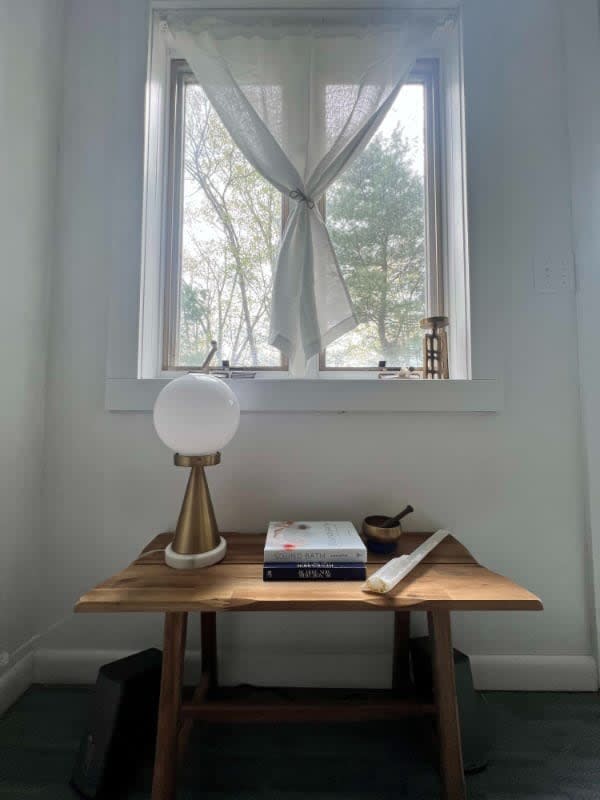 A small wooden side table with a modern globe lamp, books, and decorative items, positioned in front of a window with sheer white curtains tied back. The window offers a glimpse of trees outside.
