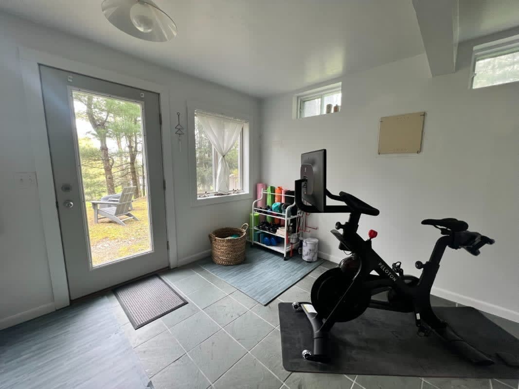 A home gym space features a Peloton exercise bike on a mat, with yoga mats and fitness accessories stored on a shelf. The room has light gray walls, dark tiled floors, and a glass door leading to the outdoors.