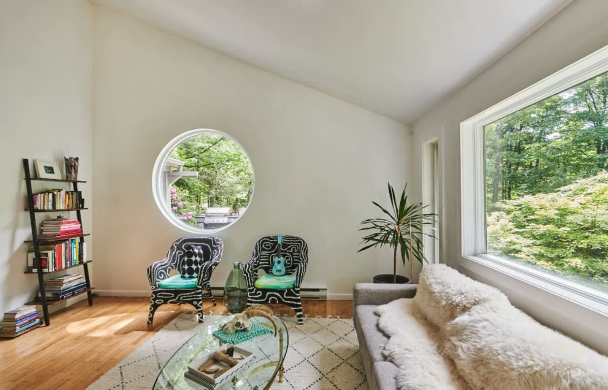 Bright living room with a unique circular window overlooking lush greenery, two patterned armchairs, and a glass coffee table on a geometric rug. A black ladder bookshelf adds a touch of eclectic style.