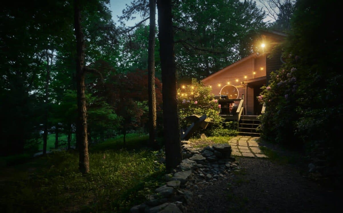 The gray-sided cabin is beautifully illuminated at dusk, with warm string lights draped around the entrance and deck area, creating a cozy ambiance. The surrounding woodland is dark, highlighting the inviting glow of the house.
