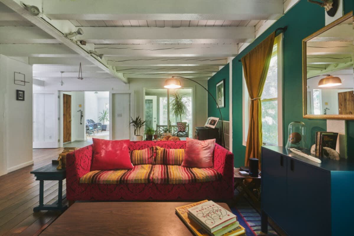 Eclectic living space with a red patterned sofa, exposed white ceiling beams, and a mix of colorful decor. A large mirror reflects the room, and a blue side table adds a pop of color.