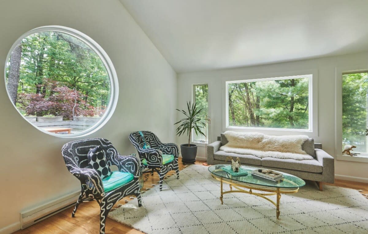 A bright living room with white walls and a vaulted ceiling, featuring a large circular window overlooking lush greenery. Two black and white wicker chairs with teal cushions face a gray sofa, centered around a glass-top coffee table on a patterned rug.