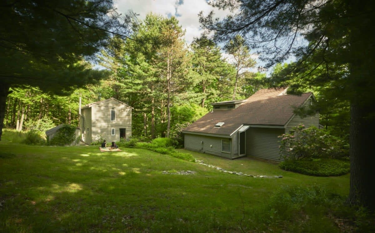 Expansive view of the property's exterior, showcasing two distinct buildings nestled within a lush, green wooded landscape. A large grassy lawn separates the main house from a smaller, taller structure.